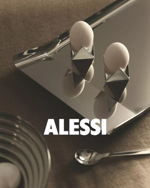 Zwei moderne, geometrische Eierbecher mit Eiern stehen auf einem reflektierenden Tablett, dazu eine Glasschüssel und ein Löffel. Unten in der Bildmitte ist in fetter Schrift „ALESSI“ zu lesen.