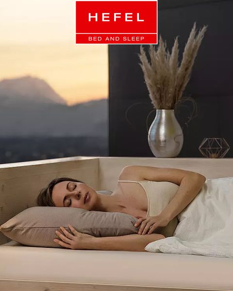 Eine Frau schläft friedlich auf der Seite in einem modernen Bett mit beigem Kissen und weißer Decke. Im Hintergrund sind eine Vase mit Pampasgras und ein Bergblick bei Sonnenuntergang zu sehen. Oben prangt das Logo von „Hefel Bed and Sleep“.
