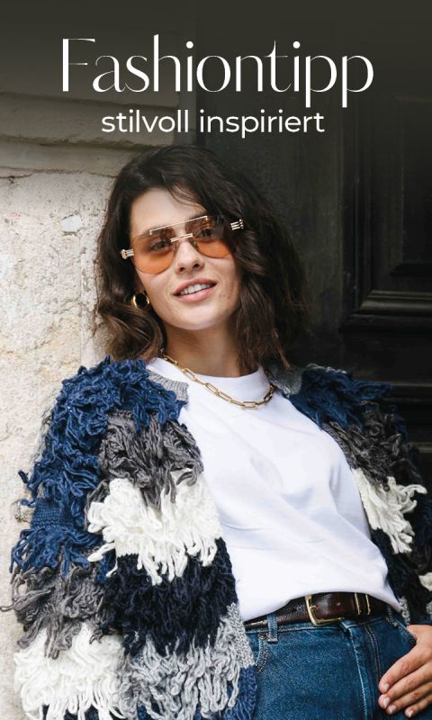 Eine Frau mit schulterlangem braunem Haar trägt eine Sonnenbrille, eine dicke Strickjacke in Blau, Weiß und Schwarz, ein weißes T-Shirt und Jeans. Sie lächelt, während sie sich gegen eine Wand lehnt. Der Text darüber lautet: "Fashiontipp stilvoll inspiriert.