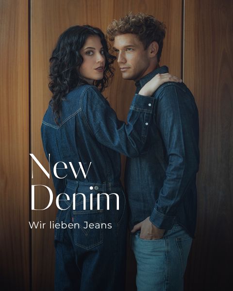 Eine Frau und ein Mann in Jeanskleidung stehen dicht beieinander vor einer Holztafel. Die Frau hat ihre Hände auf die Schultern des Mannes gelegt. Der Text lautet: "New Denim. Wir lieben Jeans.