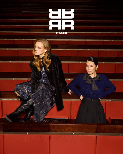 Zwei Frauen posieren stilvoll in einem Theater mit roten Sitzen. Die eine sitzt auf der Rückenlehne eines Sitzes und trägt ein gemustertes Kleid, einen schwarzen Mantel und Stiefel. Die andere steht in einem marineblauen Pullover und einem schwarzen Rock neben ihr. Über ihnen erscheint das Logo "RIANI".