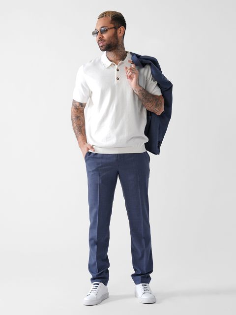 Ein Mann mit Sonnenbrille, weißem Kurzarm-Poloshirt, blauer Anzughose und weißen Turnschuhen steht vor einem schlichten hellen Hintergrund und hält einen blauen Blazer über der Schulter.