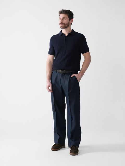 Ein Mann steht vor einem schlichten weißen Hintergrund. Er trägt ein marineblaues Poloshirt mit kurzen Ärmeln, eine dunkelblaue Hose, einen schwarzen Gürtel und braune Schuhe. Eine Hand steckt in seiner Tasche, der Blick ist zur Seite gerichtet.
