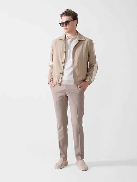 Ein Mann mit Sonnenbrille, beiger Jacke, weißem Hemd, taupefarbener Hose und beigen Slippern steht vor einem schlichten weißen Hintergrund, hat die Hände in den Taschen und blickt zur Seite.