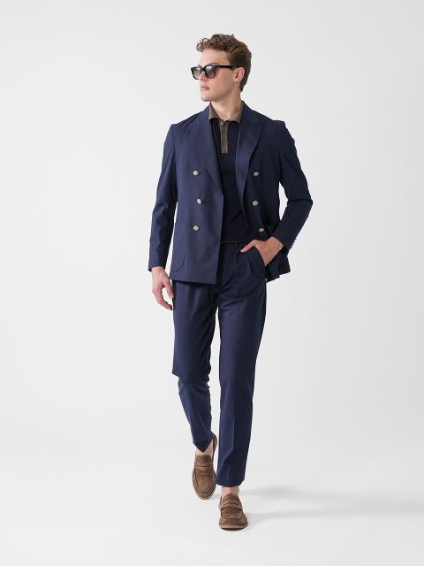 Ein Mann in einem marineblauen Zweireiheranzug, einem dunklen Hemd, braunen Slippern und einer Sonnenbrille geht selbstbewusst vor einem schlichten weißen Hintergrund.