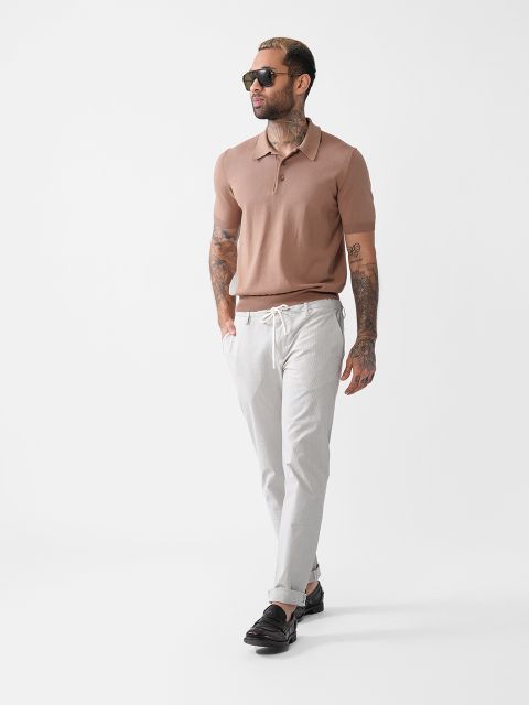 Ein Mann mit Tattoos, einer Sonnenbrille, einem hellbraunen Poloshirt mit kurzen Ärmeln, hellgrauen Hosen mit Kordelzug und schwarzen Slippern geht selbstbewusst vor einem schlichten weißen Hintergrund.