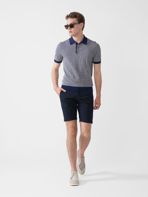 Ein Mann mit Sonnenbrille, einem kurzärmeligen Strickhemd mit Reißverschlusskragen, dunklen Shorts und hellen Slippern geht selbstbewusst vor einem schlichten weißen Hintergrund.