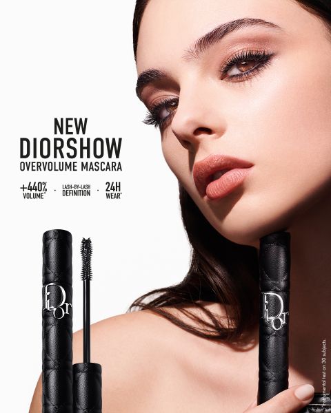 Eine Frau mit dramatischem Augen-Make-up hält eine Tube DiorShow Overvolume Mascara. Der Text hebt die Eigenschaften hervor: "+440% Volumen, Definition Wimper für Wimper, 24 Stunden Haltbarkeit." Das Mascarastäbchen ist vor ihr ausgestellt.