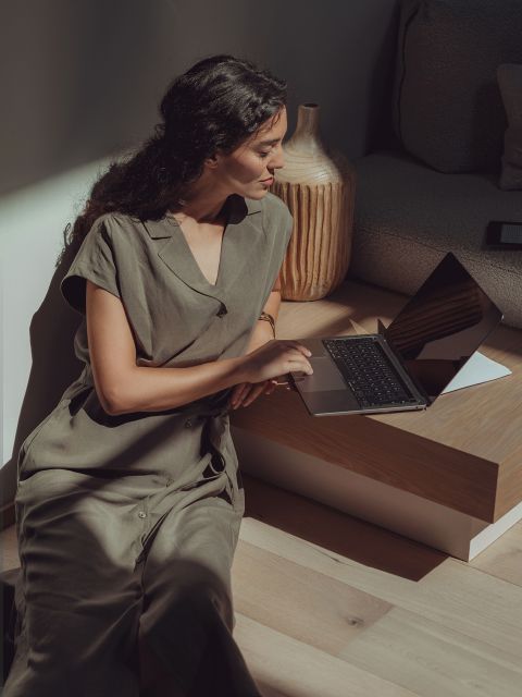 Eine Frau in einem olivgrünen Kleid sitzt auf dem Boden neben einem niedrigen Holztisch und benutzt einen Laptop. Sonnenlicht scheint auf sie, und auf dem Tisch hinter ihr steht eine große dekorative Vase.