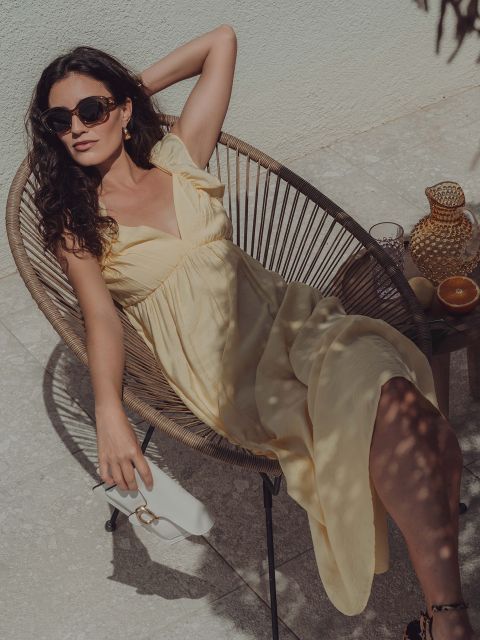 Eine Frau in einem hellgelben Kleid sitzt mit Sonnenbrille in einem runden Korbstuhl im Freien. Auf einem kleinen Tisch neben ihr stehen ein Glas, eine Karaffe und Zitrusfrüchte. Sonnenlicht wirft Schatten auf die Szene.
