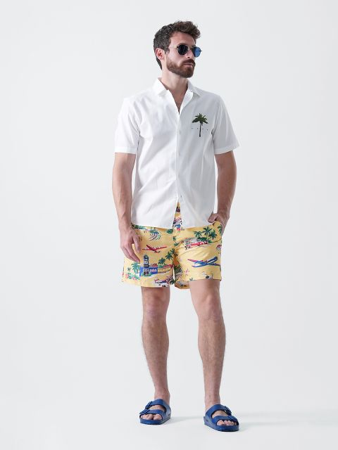 Ein Mann mit Sonnenbrille, einem weißen kurzärmeligen Hemd mit einer Palme auf der Brusttasche, gelben Shorts mit tropischem Muster und blauen Sandalen steht vor einem schlichten weißen Hintergrund.