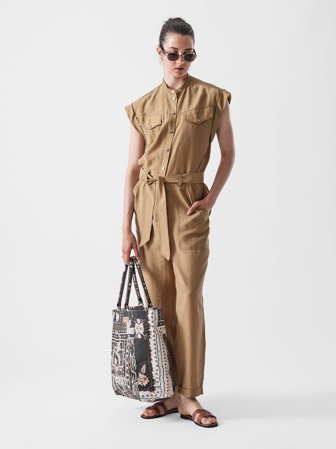Eine Person mit Sonnenbrille und einem hellbraunen Overall mit Gürtel steht vor einem weißen Hintergrund, hält eine gemusterte Einkaufstasche und trägt braune Sandalen.