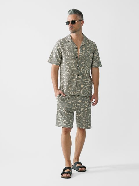Ein Mann mit kurzen grauen Haaren und Sonnenbrille trägt ein passendes gemustertes Kurzarmhemd und Shorts mit schwarzen Sandalen und steht vor einem schlichten weißen Hintergrund.