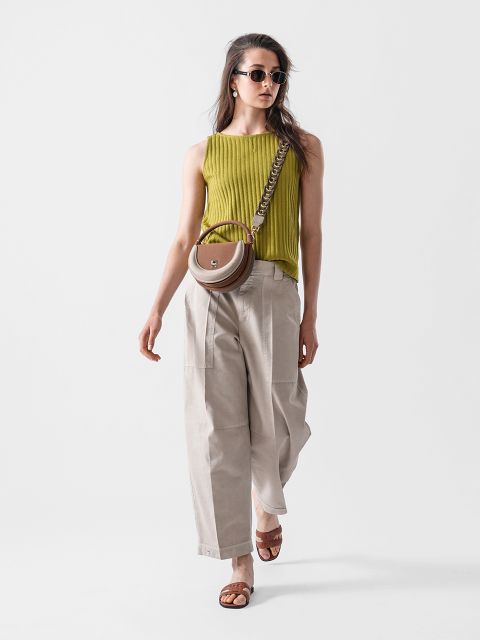 Eine Frau mit Sonnenbrille trägt ein ärmelloses limettengrünes Oberteil, weite beige Hosen und braune Sandalen. Sie trägt eine runde braune Umhängetasche mit einem klobigen Riemen und geht selbstbewusst vor einem schlichten weißen Hintergrund.