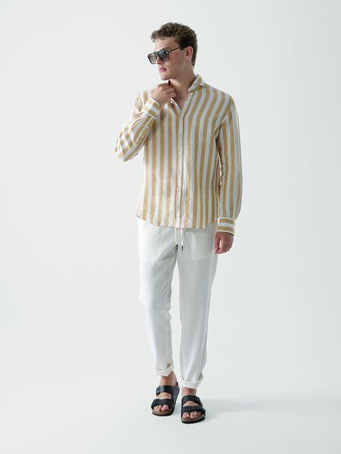 Ein Mann mit Sonnenbrille, einem beige-weißen Langarmhemd mit vertikalen Streifen, einer weißen Hose mit Kordelzug und schwarzen Sandalen steht vor einem schlichten weißen Hintergrund.