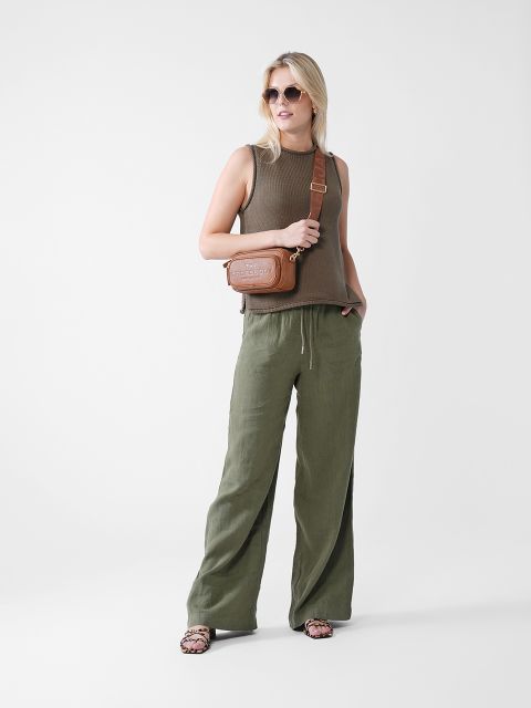 Eine Frau mit Sonnenbrille, ärmellosem braunen Top, lockeren olivgrünen Hosen und Sandalen steht mit einer Hand in der Tasche und einer braunen Umhängetasche über der Schulter vor einem schlichten weißen Hintergrund.