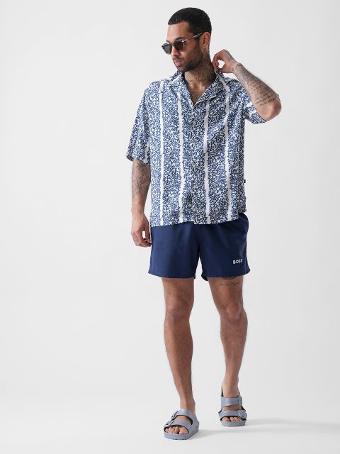 Ein Mann mit Sonnenbrille, kurzärmeligem gemustertem Hemd, marineblauen Shorts mit „BOSS“ auf dem Bein und grauen Sandalen steht vor einem schlichten hellen Hintergrund.