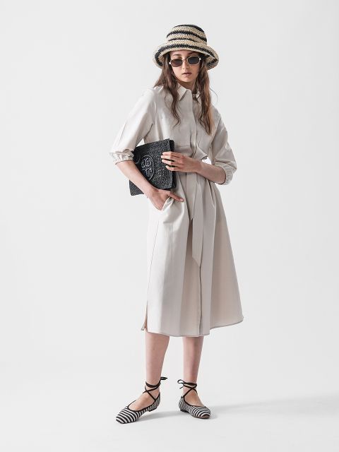 Eine Frau in einem hellen, knielangen Kleid, Strohhut und Sonnenbrille hält eine schwarze Clutch. Sie trägt schwarz-weiß gestreifte flache Schuhe und steht vor einem schlichten weißen Hintergrund.