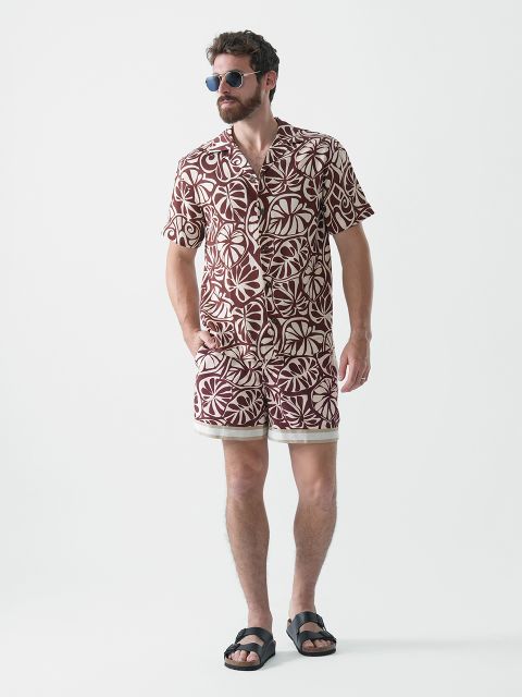 Ein Mann mit Bart und Sonnenbrille trägt ein passendes Kurzarmhemd und Shorts mit einem auffälligen braun-weißen geometrischen Muster. Er steht vor einem schlichten weißen Hintergrund und trägt schwarze Sandalen.