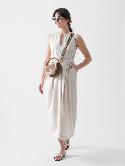 Eine Frau in einem ärmellosen weißen Midikleid mit Gürtel, einer Sonnenbrille und Sandalen sowie einer runden hellbraunen Umhängetasche mit Kettenriemen steht lächelnd vor einem schlichten weißen Hintergrund.