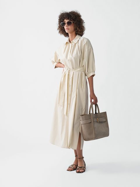 Frau mit Sonnenbrille und lockerem, cremefarbenem Midikleid mit Bindegürtel. Sie hält eine taupefarbene Handtasche in der Hand und trägt Riemchensandalen. Sie steht vor einem schlichten weißen Hintergrund.