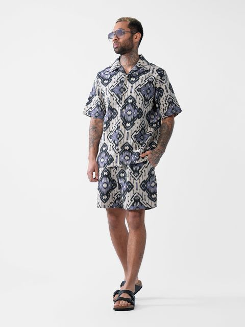 Ein Mann mit Tätowierungen, einer Sonnenbrille, einem passenden gemusterten Kurzarmhemd und Shorts sowie schwarzen Sandalen steht selbstbewusst vor einem schlichten weißen Hintergrund.