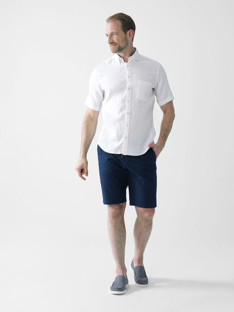 Ein Mann steht vor einem schlichten weißen Hintergrund. Er trägt ein kurzärmeliges weißes Hemd, marineblaue Shorts und graue Slipper. Er hat eine Hand in der Tasche, blickt zur Seite und lächelt leicht.