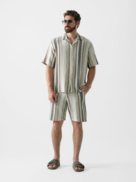 Ein Mann mit Sonnenbrille, kurzärmligem, gestreiftem Hemd mit Knopfleiste, dazu passenden gestreiften Shorts und grünen Pantoletten steht vor einem schlichten weißen Hintergrund.