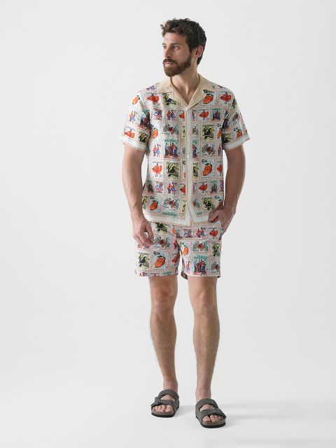 Ein Mann mit Bart steht vor einem schlichten Hintergrund. Er trägt ein passendes Kurzarmhemd, Shorts mit buntem Vintage-Print und schwarze Sandalen. Er blickt zur Seite und hat die Hände in den Taschen.