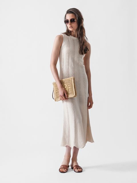 Eine Frau mit Sonnenbrille und einem ärmellosen, langen, hellbeigen Strickkleid, einer Clutch aus geflochtenem Stroh in der Hand und braunen Sandalen steht vor einem schlichten weißen Hintergrund.