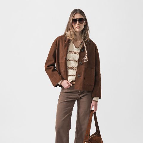 Frau mit Sonnenbrille, brauner Wildlederjacke, gemustertem Pullover und brauner Hose, die eine braune Tasche vor einem schlichten weißen Hintergrund hält.