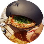 Eine Person nimmt mit einem Pizzaschieber eine frisch gebackene Pizza mit Rucola und Gemüse aus einem schwarzen Pizzaofen im Freien, der auf einem Holztisch steht.