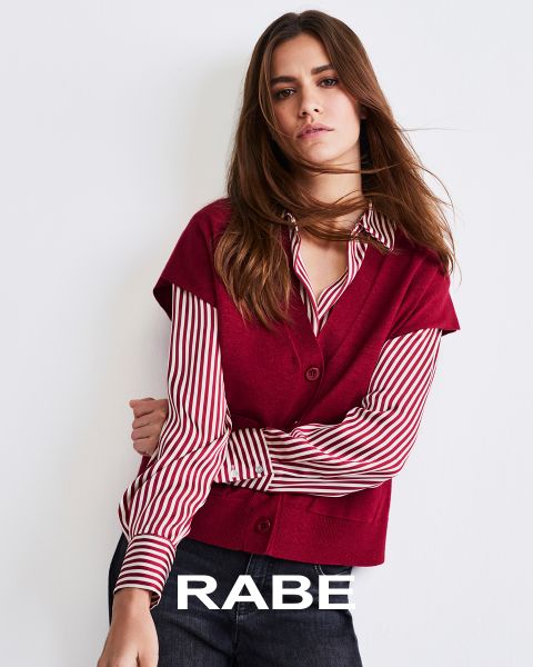 Eine Frau posiert vor einer weißen Wand und trägt eine rote ärmellose Weste über einer rot-weiß gestreiften Bluse und eine dunkle Hose. Am unteren Rand des Bildes ist das Wort "RABE" zu lesen.