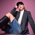 Eine Person sitzt vor einem rosafarbenen Hintergrund, trägt einen Nadelstreifenblazer, eine weiße Bluse, Jeansshorts, hohe schwarze Stiefel und eine schwarze Kappe, mit einem selbstbewussten Gesichtsausdruck und einer entspannten Pose.
