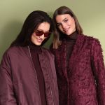 Zwei Frauen stehen Seite an Seite vor einem hellgrünen Hintergrund. Die eine trägt eine Sonnenbrille und eine weinrote Jacke, die andere einen strukturierten weinroten Blazer. Beide haben dunkles Haar und lächeln.