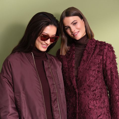 Zwei Frauen stehen Seite an Seite vor einem hellgrünen Hintergrund. Die eine trägt eine Sonnenbrille und eine weinrote Jacke, die andere einen strukturierten weinroten Blazer. Beide haben dunkles Haar und lächeln.