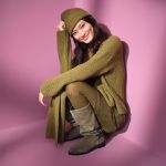 Eine Frau sitzt lächelnd vor einem rosafarbenen Hintergrund und trägt einen olivgrünen Strickpullover, eine passende Mütze, einen Schal, eine Hose und graue Wildlederstiefel.