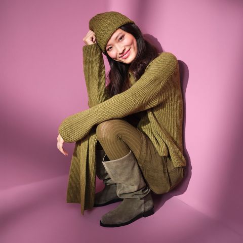 Eine Frau sitzt lächelnd vor einem rosafarbenen Hintergrund und trägt einen olivgrünen Strickpullover, eine passende Mütze, einen Schal, eine Hose und graue Wildlederstiefel.