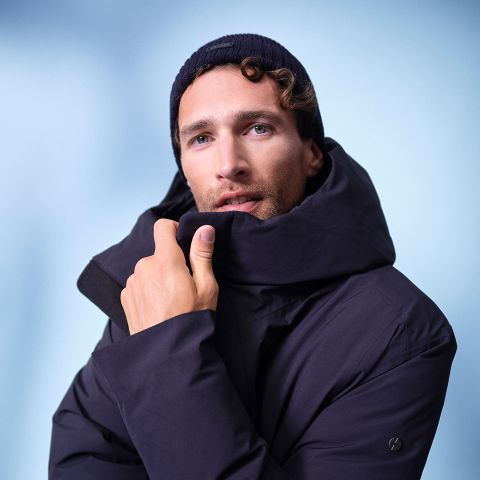 Ein Mann, der einen dunklen Wintermantel und eine dunkle Strickmütze trägt, blickt in die Kamera und hält sich den Kragen seines Mantels vor einem weichen blauen Hintergrund vors Gesicht.