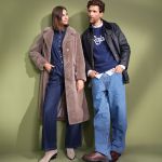 Eine Frau in einem langen Mantel aus Kunstpelz und einem Jeans-Outfit steht neben einem Mann in Lederjacke, blauem "Ralph"-Pullover und Baggy Jeans. Beide sehen vor einem gedämpften grünen Hintergrund stilvoll aus.