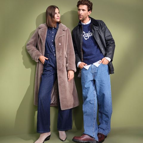 Eine Frau in einem langen Mantel aus Kunstpelz und einem Jeans-Outfit steht neben einem Mann in Lederjacke, blauem "Ralph"-Pullover und Baggy Jeans. Beide sehen vor einem gedämpften grünen Hintergrund stilvoll aus.