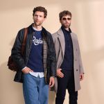 Zwei Männer posieren vor einem beigen Hintergrund; einer trägt eine schwarze Jacke, blaue Jeans und einen marineblauen Pullover mit der Aufschrift "Ralph", der andere einen grauen Mantel, eine dunkle Hose und eine Sonnenbrille.