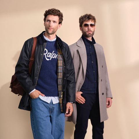 Zwei Männer posieren vor einem beigen Hintergrund; einer trägt eine schwarze Jacke, blaue Jeans und einen marineblauen Pullover mit der Aufschrift "Ralph", der andere einen grauen Mantel, eine dunkle Hose und eine Sonnenbrille.