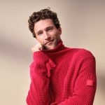 Ein Mann mit lockigem braunem Haar und blauen Augen, der einen strukturierten roten Rollkragenpullover trägt, lächelt sanft, während er sein Kinn auf seine Hand vor einem beigen Hintergrund stützt.