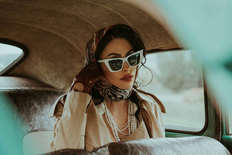 Eine elegante Frau mit weißer Sonnenbrille, Kopftuch, Perlen, Handschuhen und einem beigen Trenchcoat sitzt auf dem Rücksitz eines Oldtimers und schaut aus dem Fenster.