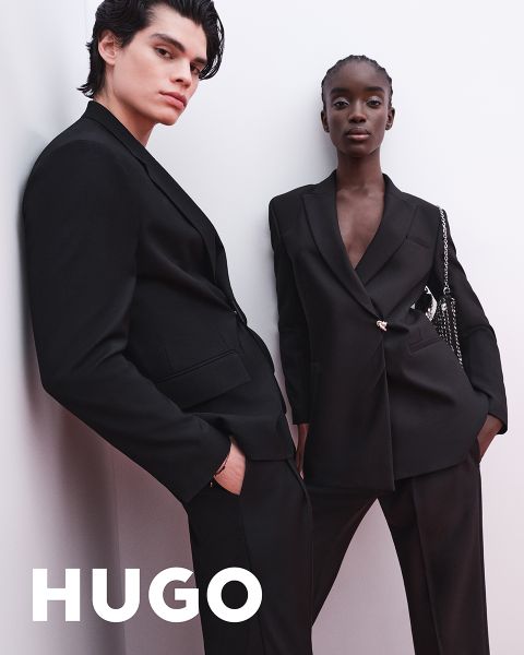 Zwei Models in eleganten schwarzen Anzügen posieren selbstbewusst vor einem schlichten weißen Hintergrund. Das Wort "HUGO" erscheint in fetten weißen Buchstaben in der unteren linken Ecke und weist auf eine Werbung für eine Modemarke hin.