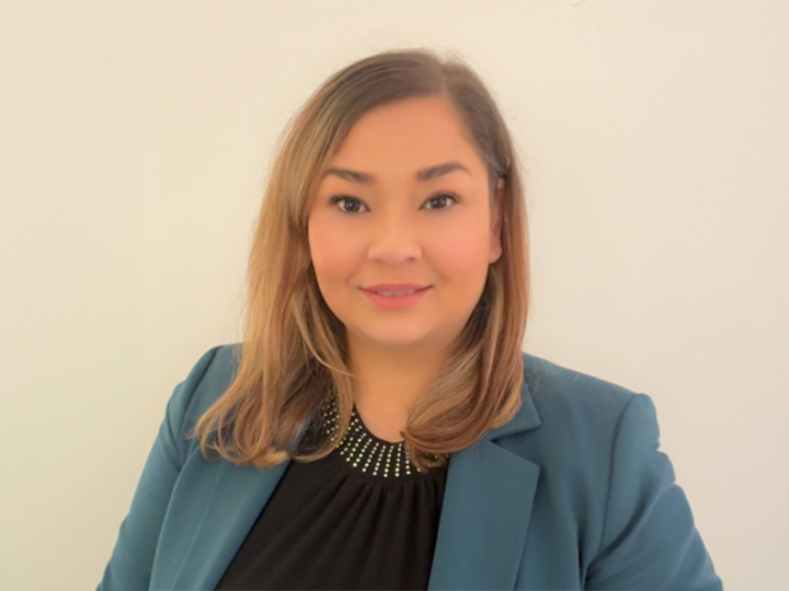 Frau mit schulterlangem hellbraunem Haar, die einen blaugrünen Blazer und ein schwarzes Oberteil trägt, lächelt vor einem schlichten hellen Hintergrund.