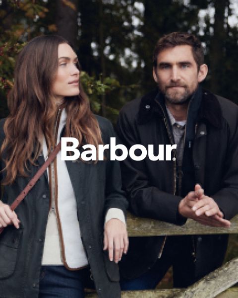 Eine Frau und ein Mann in Barbour-Jacken stehen im Freien an einem Holzzaun, mit Bäumen im Hintergrund. Das Barbour-Logo ist in der Mitte des Bildes in weißer Farbe hervorgehoben.