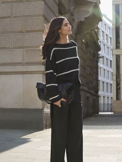 Eine Frau mit langen Haaren steht auf einem Bürgersteig in der Stadt. Sie trägt einen schwarzen Pullover mit weißen Streifen und eine schwarze Hose mit weitem Bein und hält eine große schwarze Clutch in der Hand, während sie von hinten vom Sonnenlicht angestrahlt wird.