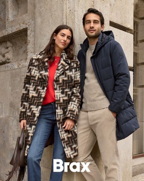 Eine Frau in einem gemusterten Mantel und einem roten Pullover steht neben einem Mann in einem marineblauen Wintermantel und einer beigen Hose. Sie lehnen an einer Steinmauer im Freien. Das Wort "Brax" erscheint am unteren Rand des Bildes.